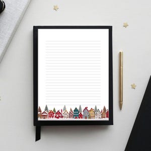 May include: A lined notepad with a black frame and a decorative border of colorful houses. A gold pen and a black notebook are next to the notepad. The scene is set on a white surface with gold star-shaped confetti.