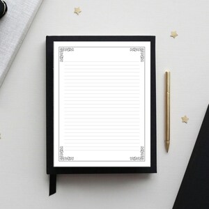 Op de afbeelding: Een gelijnd notitieblok met decoratieve hoekontwerpen ligt in een zwart frame op een wit oppervlak. Een gouden pen en een zwart notitieboek liggen in de buurt. Het notitieblok is open, klaar om te schrijven.