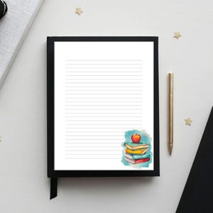 May include: A lined notepad with a black frame and a watercolor illustration of a stack of books and an apple. A gold pen rests beside the notepad. The scene is set on a white surface with decorative stars and a black ribbon.