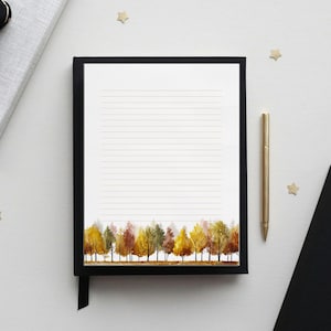 May include: A lined notepad with a black frame and a watercolor forest design at the bottom. A gold pen rests beside the notepad. A black notebook and a gift box are also in the image. Gold star-shaped confetti is scattered around.