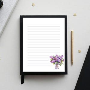 Peut inclure: Un bloc-notes ligné avec un cadre noir, orné d'une illustration à l'aquarelle de lilas violets dans un vase en verre. Un stylo doré est posé à côté du bloc-notes. Le bloc-notes est sur une surface blanche avec un livre noir et un nœud décoratif.