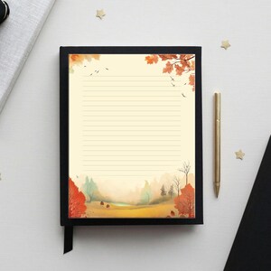 May include: A lined notepad with a black border and a watercolor autumn landscape scene. The notepad is open, with a gold pen resting beside it. A black ribbon is attached to the notepad.