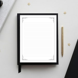 May include: A lined notepad with decorative corner accents sits in a black cover with a ribbon bookmark. A gold pen rests beside the notepad. The scene is set on a white surface with a few gold star-shaped decorations.