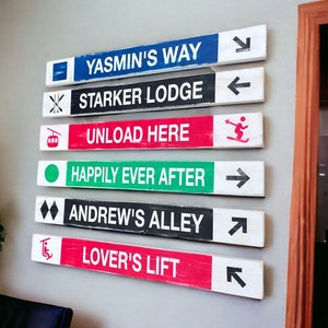 May include: A set of seven wooden signs with white painted text and arrows. The signs are painted in different colors, including red, green, black, and blue. The signs read: Yasmin's Way, Starker Lodge, Unload Here, Happily Ever After, Andrew's Alley, and Lover's Lift.