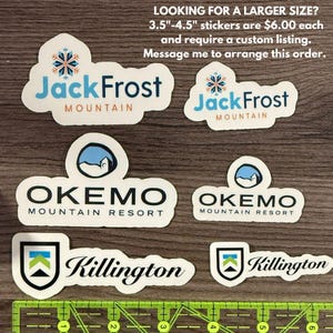 May include: Six different ski resort stickers on a wooden surface. The stickers are white with black text and logos. The logos include Jack Frost Mountain, Okemo Mountain Resort, and Killington.
