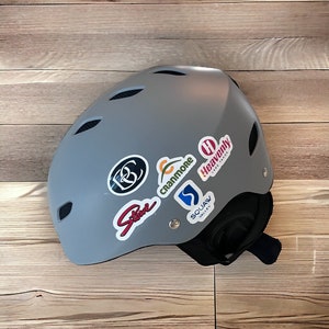 May include: Gray ski helmet with various colorful ski resort stickers, including "Heavenly Lake Tahoe", "Squaw Valley", "Cranmore", and "Stowe".