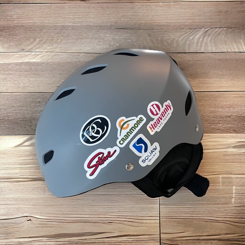 May include: Gray ski helmet with various colorful ski resort stickers, including "Heavenly Lake Tahoe", "Squaw Valley", "Cranmore", and "Stowe".