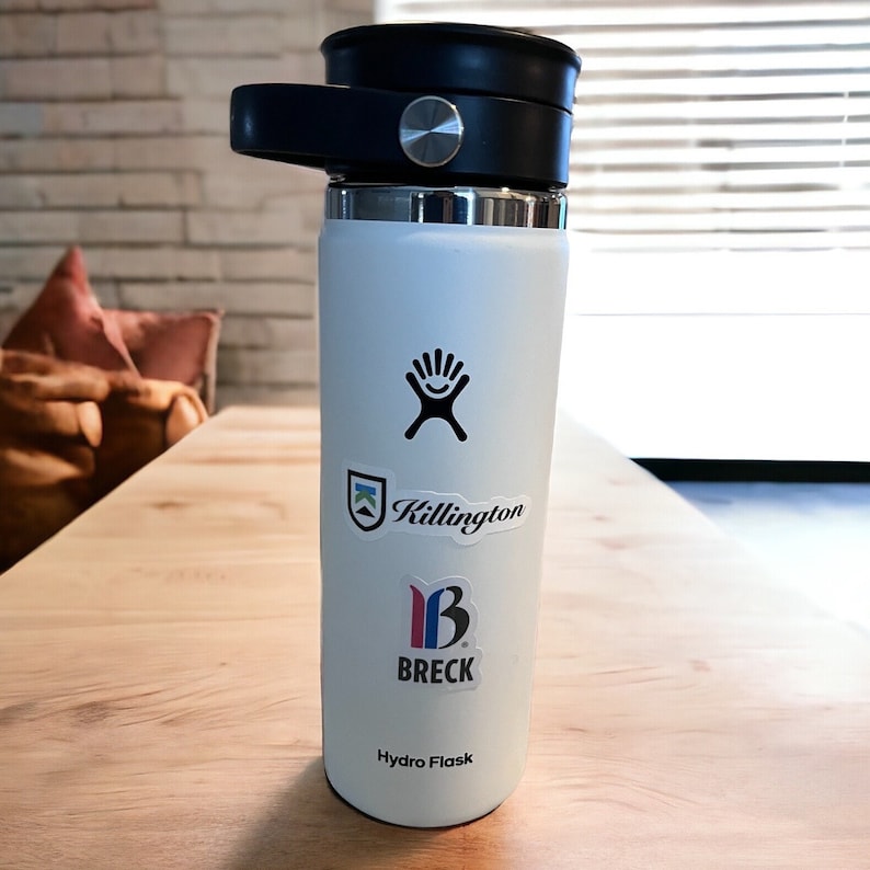 May include: White Hydro Flask water bottle with a black lid and a black hand logo. The bottle has three stickers on it: a Killington ski resort logo, a Breck ski resort logo, and a Hydro Flask logo.