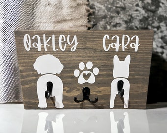 May include: A wooden dog leash holder with two hooks. The holder has the names "Oakley" and "Cara" written on it, and a paw print with a heart in the middle.