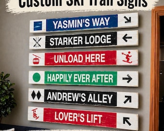 May include: A collection of custom ski trail signs with various names and directional arrows. The signs are rectangular, with a distressed wood appearance and colorful backgrounds. Text includes "YASMIN'S WAY," "STARKER LODGE," "UNLOAD HERE," "HAPPILY EVER AFTER," "ANDREW'S ALLEY," and "LOVER'S LIFT." Bike trails are also available.