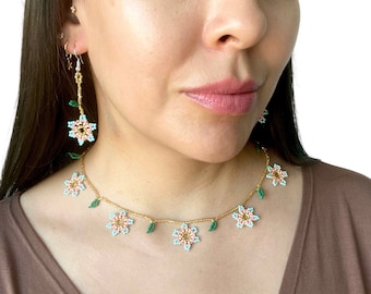 May include: A necklace and earring set made of beads. The necklace and earrings feature flower-shaped beads in pink, blue, and gold, with green leaf beads. The necklace and earrings are on a gold-coloured chain.