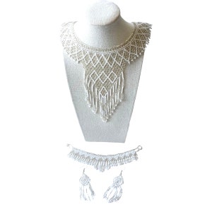 May include: A white beaded necklace, bracelet, and earring set. The necklace is displayed on a mannequin and features a bib design with a diamond pattern and fringe. The bracelet and earrings have matching beaded details.