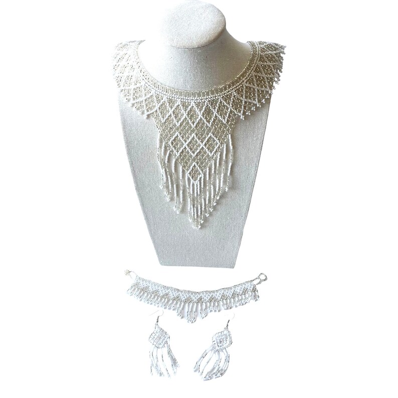 May include: A white beaded necklace, bracelet, and earring set. The necklace is displayed on a mannequin and features a bib design with a diamond pattern and fringe. The bracelet and earrings have matching beaded details.