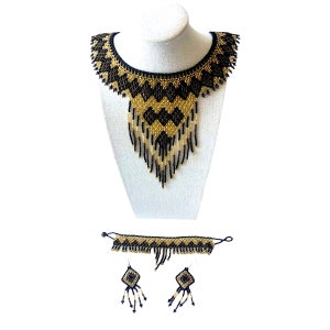May include: A black and gold beaded necklace, bracelet, and earring set. The necklace is displayed on a white mannequin bust. The jewelry features a geometric diamond pattern and fringe detailing. The set includes a choker-style bracelet and dangling earrings.