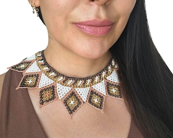 May include: A beaded collar necklace with a geometric design. The necklace features white, brown, and gold beads, with pink and black accents. The necklace is worn around the neck.