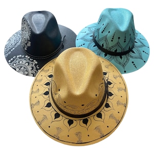 May include: Three fedora hats with mandala designs. One is black with a white mandala, another is light blue with a black mandala, and the third is tan with a black mandala. Each hat has a black band around the crown.