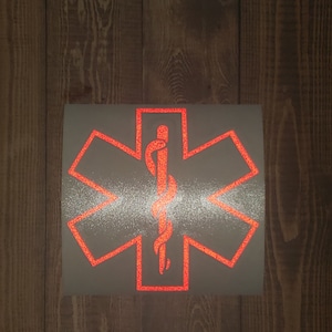 Peut inclure: Un autocollant rouge en forme d'étoile de la vie sur fond argenté pailleté. L'étoile de la vie est un symbole utilisé par les services médicaux d'urgence.