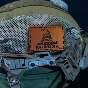 May include: A tan and green camouflage helmet with a brown leather patch that says "Don't Tread On Me" and a snake design.