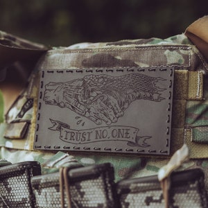 May include: A black leather patch with a skeletal hand shake design and the text "TRUST NO ONE". The patch is attached to a camouflage tactical vest with multiple magazine pouches.