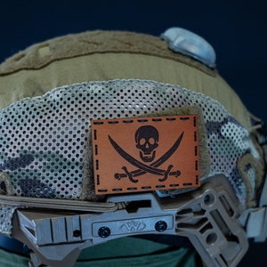 May include: A tan and green camouflage helmet with a brown leather patch featuring a skull and crossbones. The patch has the words "Jolly Roger" stitched on it.
