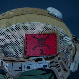May include: A camouflage helmet with a red patch featuring a skull and crossbones. The helmet has a mesh panel and a side-mounted accessory.