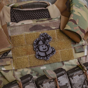 May include: A camouflage tactical vest with a black patch featuring a stick figure wearing a helmet and holding binoculars. The vest has multiple magazine pouches with black magazines inserted.