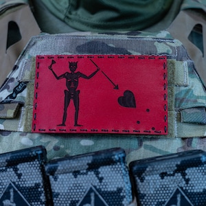 May include: A red patch with a black skull and crossbones design, a heart, and three dots. The patch is sewn onto a camouflage tactical vest with black magazine pouches.