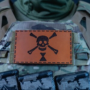 May include: A brown leather patch with a black skull and crossbones design. The patch is attached to a camouflage tactical vest with multiple magazine pouches.