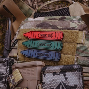 May include: Three crayon-shaped patches in red, green, and blue, each with the text "EMERGENCY SNACK" in a white, rounded font. The patches are affixed to a tactical vest with a camouflage pattern. Additional tactical gear is visible.