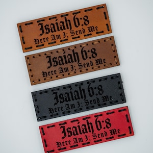 May include: Four rectangular leather patches in shades of brown, black, and red. Each patch has the text "Isaiah 6:8 Here Am I; Send Me" stitched in black. The patches are arranged on a white surface.