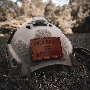 May include: A tan helmet with a brown leather patch that says "VIOLENCE = RESULTS" on it. The helmet is lying in green grass.