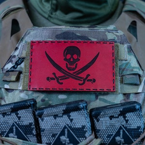 May include: A red patch with a black skull and crossbones design sewn onto a camouflage tactical vest. The vest has multiple magazine pouches and the text "MULTICAM" is visible on the bottom.