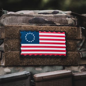 May include: A rectangular patch featuring the Betsy Ross flag design, with a blue field of white stars and red and white stripes. The patch is sewn onto a camouflage fabric, with a dark green background.