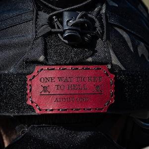 May include: A red leather patch with the text "One Way Ticket To Hell" and two skull and crossbones symbols. The text "Admit One" is printed below the main text.