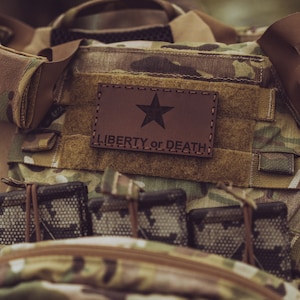 May include: A brown leather patch with a star and the text "LIBERTY OR DEATH" sewn onto a camouflage tactical vest.