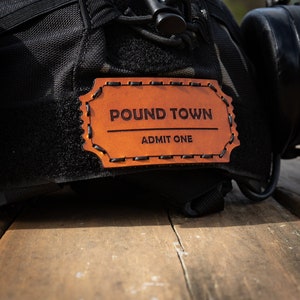 May include: A brown leather patch with the text "POUND TOWN" and "ADMIT ONE" stitched on it. The patch is attached to a black tactical helmet.