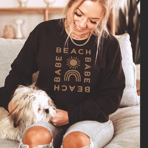Puede incluir: Una sudadera negra con un dise&ntilde;o gr&aacute;fico marr&oacute;n que dice "Beach Babe" con un sol y un arco&iacute;ris. La sudadera la lleva puesta una persona con el pelo largo y rubio.