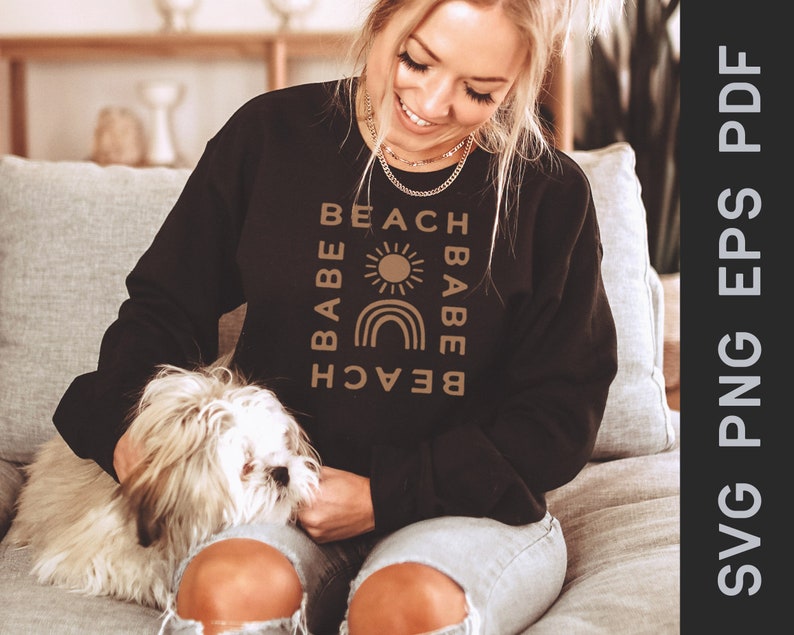 Puede incluir: Una sudadera negra con un dise&ntilde;o gr&aacute;fico marr&oacute;n que dice "Beach Babe" con un sol y un arco&iacute;ris. La sudadera la lleva puesta una persona con el pelo largo y rubio.