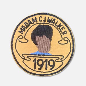 May include: A yellow circular patch with black embroidery. The patch features a portrait of a woman with short dark hair, a blue shirt, and the text "MADAM C.J. WALKER 1919".