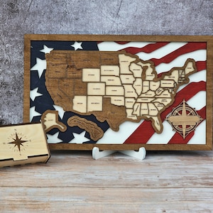 May include: Wooden United States map with state outlines, set against a red, white, and blue American flag design. Includes a small wooden box with a compass rose design. The map is displayed on a wooden stand.