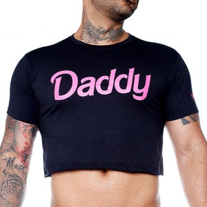 May include: A black crop top with the word "Daddy" printed in pink across the front. The shirt has short sleeves and a crew neck. The text is in a cursive font.