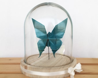 Könnte beinhalten: Ein türkisfarbener Origami-Schmetterling, der unter einer Glasglocke auf einem Holzsockel mit einem weißen Band präsentiert wird.