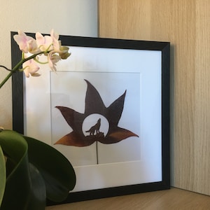 Może przedstawiać: Wydruk w ramie przedstawiający brązowy liść z sylwetką wilka w środku. Liść ma kształt gwiazdy, a wilk wyje do księżyca. Wydruk znajduje się w czarnej ramie i wisi na ścianie.