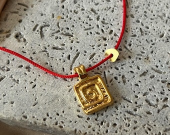 May include: A gold-colored pendant necklace with a red cord. The pendant is square with an engraved spiral design. The necklace is displayed on a textured gray surface.