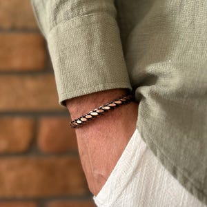 K&ouml;nnte beinhalten: Ein kupferfarbenes geflochtenes Armband wird am Handgelenk getragen, mit einem gr&uuml;nen Langarmhemd und einer wei&szlig;en Hose im Hintergrund. Das Armband besteht aus ineinander verwobenem Kupfer und bronzefarbenem Metall.