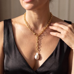 May include: A gold-toned chain necklace featuring a Y-shaped design with a large, irregular white baroque pearl pendant suspended from the centre. The necklace consists of thick, interlocking oval links with a matte finish and a secure clasp.