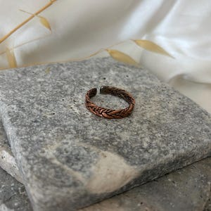 Könnte beinhalten: Ein kupferfarbener Metallring mit einer komplexen geflochtenen oder gewebten Textur auf dem gesamten Band. Der Ring hat ein offenes Design mit einer kleinen Lücke an der Oberseite und zeigt ein dunkles, oxidiertes Finish in den Vertiefungen.