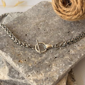 Puede incluir: Un collar de cadena de metal plateado con un cierre de palanca. El cierre consta de un anillo circular sostenido por un elemento de mano esculpido y una barra de metal recta que pasa a través del anillo para asegurar los eslabones.