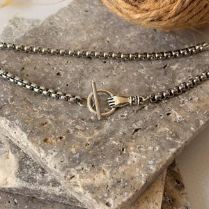 May include: A silver-toned box chain necklace featuring a toggle clasp. The clasp mechanism consists of a circular ring and a T-bar, with one side of the ring designed to resemble a small hand grasping the metal bar to secure the necklace closure.