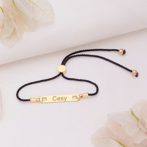 Puede incluir: Un brazalete ajustable de cordón negro con un dije de barra dorada grabado con el nombre "Cesy".